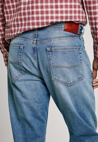 Light blue denim jeans with two back pockets, contrast stitching, and a brown leather logo patch at the waistband. Textured finish.