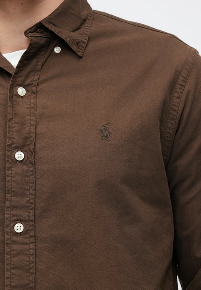 Camisa marrón de botones con botones blancos, llevada sobre una camiseta blanca, que presenta un pequeño logo bordado de un jugador de polo en el pecho.
