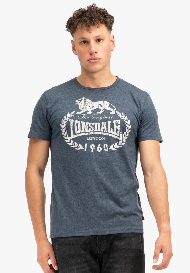 T-shirt de algodón azul oscuro con un gráfico vintage blanco que presenta un león y el texto "The Original Lonsdale London 1960" en el frente.