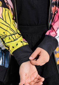 Kleurrijke bomberjack met patroon, voorzien van elastische zwarte manchetten en rits, met ontwerpen in geel, roze en blauw tegen een donkere achtergrond.