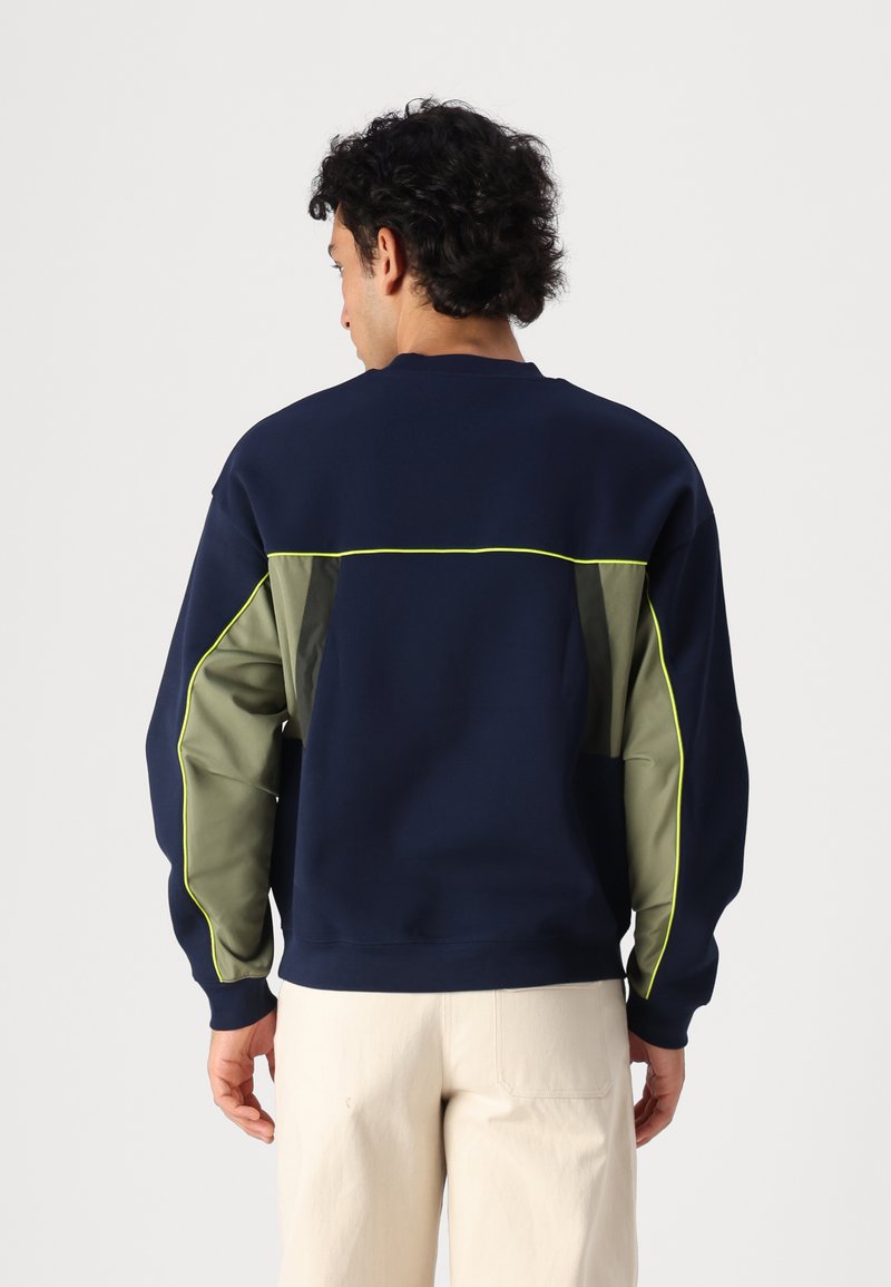 Sweat-shirt marine avec des accents vert olive, des bordures néon et une coupe décontractée. Tissu doux et poignets côtelés. Design en patchwork au dos.