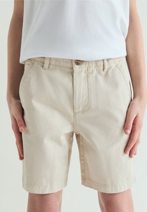 Shorts décontractés beige portés par une personne les mains reposant sur les côtés, associés à une chemise blanche rentrée.