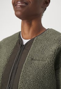 Veste vert olive avec un tissu en polaire texturé d'un côté et un panneau lisse de l'autre, avec une fermeture zippée et un logo en relief.