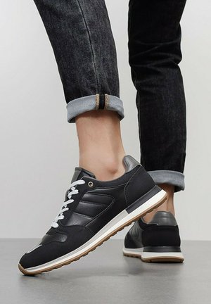 Personne portant des baskets basses noires avec des lacets blancs et des semelles en gomme, associées à un jean denim foncé retroussé, sur un sol gris.