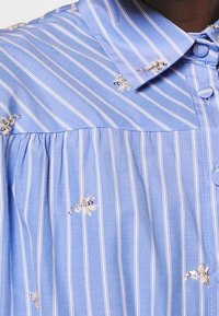 Ljusblå skjorta med vertikala vita ränder, med broderade blommönster och klassisk krage, tillverkad av en mjuk bomullsblandning.