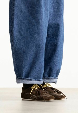 Persoon met opgerolde blauwe denim jeans en donkerbruine sneakers met niet-overeenkomende gele en bruine veters staand op een lichte vloer.