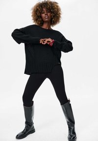 Pull en tricot noir avec ourlet côtelé et manches longues, associé à un legging noir et des bottes noires brillantes jusqu'aux genoux. Accent avec un bracelet rouge.