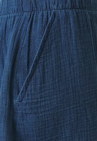 Gilinys audinys tamsiai mėlynos spalvos, su tekstūruotu, susiraukšlėjusių paviršiumi. Yra elastingas juosmuo ir šoninės kišenės detalė.