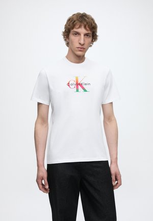 Νέος άνδρας με σγουρά μαλλιά που φοράει ένα λευκό T-shirt Calvin Klein με πολύχρωμο λογότυπο, σε συνδυασμό με μαύρο παντελόνι, στέκεται μπροστά σε λευκό φόντο.
