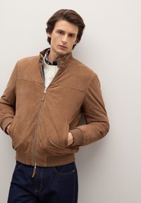 Braune Wildleder-Bomberjacke mit Reißverschluss vorne, hohem Kragen und gerippten Bündchen. Getragen über einem cremefarbenen Zopfmusterpullover und dunklen Jeans.