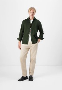 Mørkegrønn skjorte med knapper, hvit t-skjorte under, beige bukser og svarte sko. Jevn stoff, kragedesign, avslappet passform.