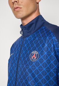 Giacca blu con zip caratterizzata da un motivo geometrico in toni di blu scuro e una patch con il logo del Paris Saint-Germain sul petto.
