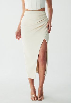 Jupe midi couleur crème avec une fente sur le côté, design drapé, texture douce et tissu lisse. Portée avec des sandales à talons transparentes.