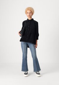 Svart skjorte med knappemønster, kombinert med blå utsvingte jeans og svarte joggesko. Modellen holder en liten svart veske.