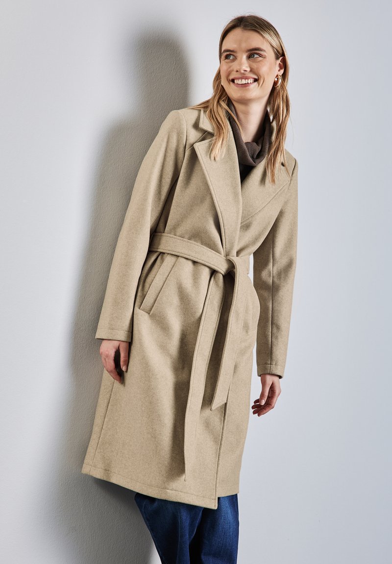 Beżowy wełniany płaszcz z paskiem w talii, klapami, długimi rękawami i bocznymi kieszeniami, zestawiony z niebieskimi dżinsami na neutralnym tle.