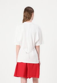 Witte top met korte mouwen en een relaxte pasvorm, met een strikdetail op de rug en een lichte glans. Gecombineerd met rode shorts, wat een contrast creëert.