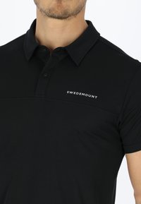 Svart polo skjorta i texturerad väv, med knäppt krage och en horisontell dekorativ linje. "SWEDMOUNT"-logotyp i vitt.