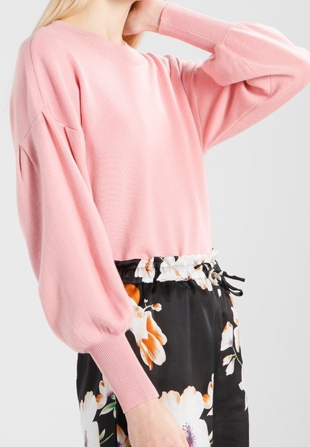 Femme portant un pull à manches longues rose pâle associé à un pantalon noir à motifs floraux avec une taille à cordon.