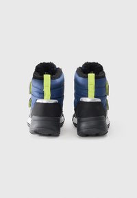 Une paire de bottes d'hiver isolées bleues avec des semelles noires, des poignées néon jaunes et une doublure en polaire noire, vues de l'arrière sur un fond blanc.