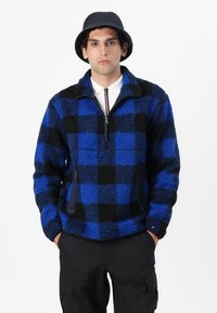 Blå- og sortternet fleecepullover med lynlås ved halsen, to sidelommer og et struktureret stof. Sammen med sorte cargo bukser og en bucket hat.