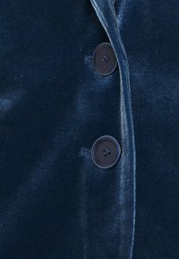Gros plan sur une veste en velours bleu marine avec deux boutons noirs ronds, une texture lisse et un léger éclat, dotée d'un col châle.