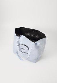 Sac fourre-tout en toile bleu clair avec intérieur noir, arborant le logo "Karl Lagerfeld Paris" et une poche zippée à l'intérieur. Texture douce, forme structurée.