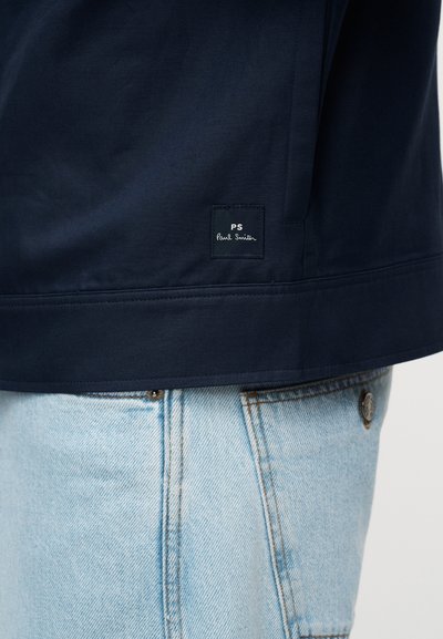 Πλέγμα ναυτικού μπουφάν με μικρό μαύρο ετικέτα "PS Paul Smith" πάνω από την τσέπη ανοιχτό μπλε τζιν.