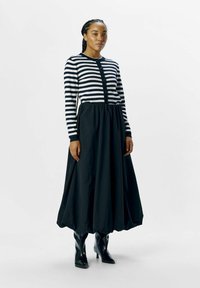 Cardigan à rayures noires et blanches à manches longues avec des boutons, associé à une jupe noire fluide. Des bottes noires à talons montantes complètent le look.