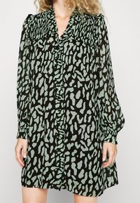 Mustapuku, jossa on vihreä abstrakti leopardi-kuosi, napit edessä, röyhelökaulus ja pitkät hihat joustavuusnauhoilla. Kevyt, virtaava kangas.