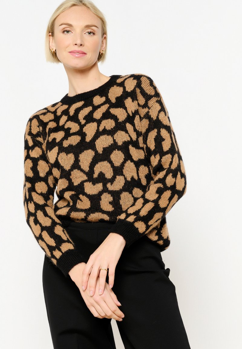 Leopardenmuster-Pullover in Schwarz und Beige. Weiches Strickmaterial mit Rundhals und langen Ärmeln. Gerippte Bündchen und Saum. Modell steht mit gefalteten Händen.