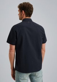 Meri pendek navy dengan kain tekstur, hem lurus, dan detail kancing belakang yang halus. Dikenakan dengan kerah klasik dan permukaan yang halus.