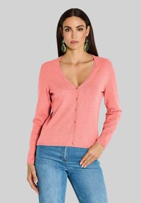 Koraalkleurige V-hals cardigan met lange mouwen, voorzien van een knoopsluiting en geribbelde onderrand. Gepaard met blauwe jeans, op een grijze achtergrond.