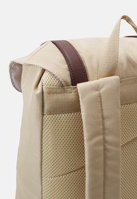 Sac à dos en tissu beige avec une sangle en cuir brun, présentant un panneau latéral en maille texturée et une fermeture enroulée.