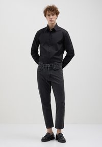 Camicia nera con bottoni e maniche lunghe, abbinata a jeans grigi scuri a sigaretta e mocassini neri, creando un look casual moderno.
