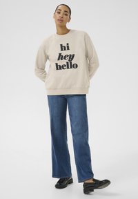 Beige mikina s výrazným černým textem "hi hey hello", kombinovaná s modrými širokými džíny a černými mokasínami, postavená před jednobarevným pozadím.