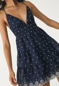 Granatowa sukienka z głębokim dekoltem w kształcie litery V, regulowanymi ramiączkami, wariantową konstrukcją, haftowanymi białymi wzorami przypominającymi gwiazdy oraz koronkowym wykończeniem na dole.