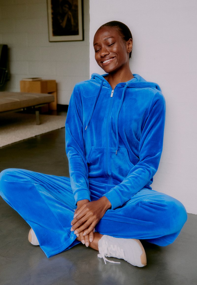 Kék bársony szabadidőruha kapucnival, elöl cipzárral és zsebekkel. A modell keresztbe tett lábakkal ül, fehér tornacipőt visel, sima padlón.