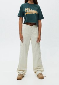 Gröna avklippta t-shirts med "Chicago" text i orange; beige vida cargo-byxor med fickor; i kombination med bruna sneakers.