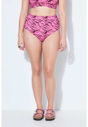 Frau trägt hochgeschnittene pinkfarbene Bikinihosen mit Zebramuster, passendes Oberteil, mehrere Ringe und pinke Sandalen mit Schnallen.