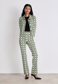 Grüner Anzug mit schwarzen Akzenten; taillierte Jacke mit geometrischem Muster, passende Hose und schwarze High Heels. Strukturiertes Gewebe.