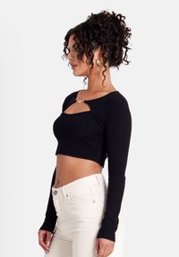 Schwarzes, kurz geschnittenes Oberteil mit langen Ärmeln, runder Ausschnittform und einem Schlüsselloch-Ausschnitt, kombiniert mit hochtaillierten cremefarbenen Hosen.