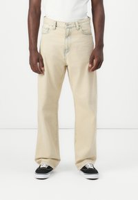 Kaki-fargede vide jeans laget av denim, med knappestolpe, fem-lomme design og subtile sømdetaljer. Sorte joggesko synlige.