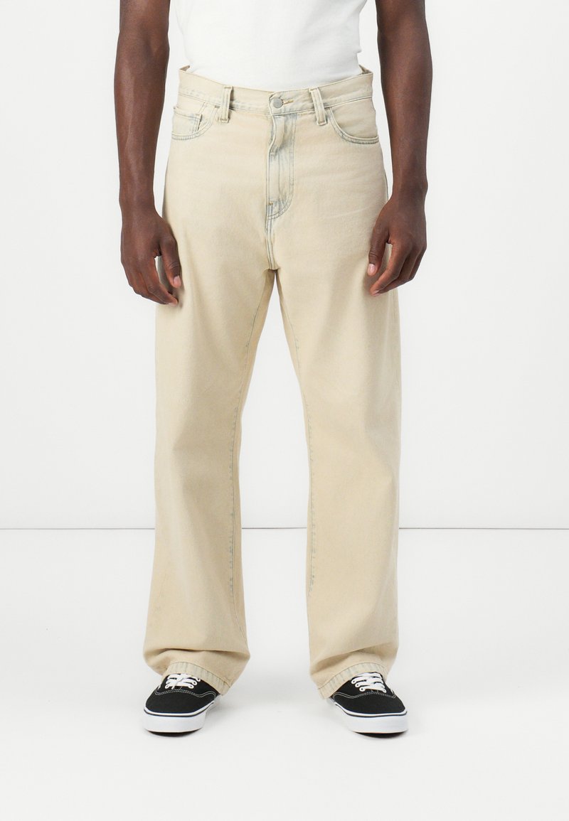 Kaki-fargede vide jeans laget av denim, med knappestolpe, fem-lomme design og subtile sømdetaljer. Sorte joggesko synlige.