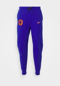 Lila Jogginghosen aus weichem Material mit einem orangefarbenen Löwenemblem und dem Nike-Logo, mit einem Kordelzugbund und gerippten Bündchen.