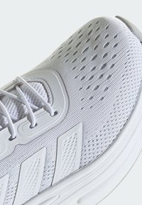 Bijela atletska cipela s mrežastim gornjim dijelom, teksturiranim bočnim naglascima i ublaženim potplatima. Ima tanke vezice i otvore za ventilaciju radi prozračnosti.