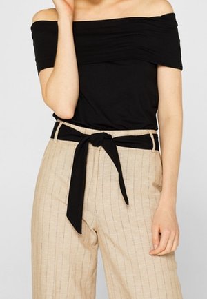 Kvinde iført en sort off-shoulder-top, beige højtaljede bukser med lodrette striber og et sort bælte bundet i en sløjfe.