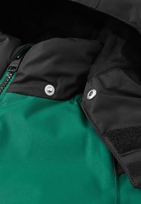 Zwarte en groene jas met een ritssluiting, drukknoopsluitingen en klittenbandaccenten. De stof is glad met een matte afwerking.