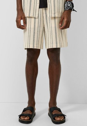 Mann trägt beige gestreifte Shorts, schwarze Sandalen und ein schwarzes Bandana, das um sein Handgelenk gebunden ist, und steht vor einem einfarbigen hellen Hintergrund.