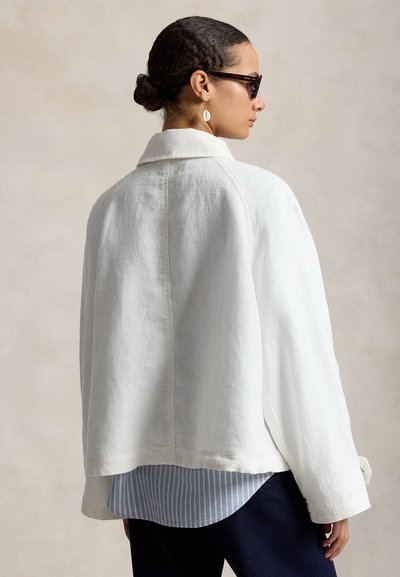 Femeie cu părul prins în coc poartă ochelari de soare închiși la culoare, geacă albă oversized, cămașă albastră cu dungi și cercei cu scoică atârnată, privirea pe profil.