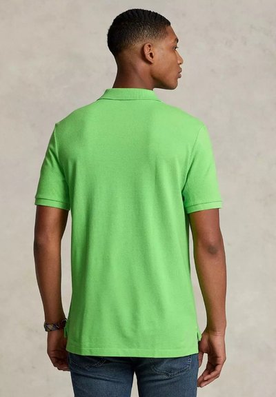 Giovane uomo con capelli corti che indossa una polo verde brillante, di spalle, in piedi contro uno sfondo neutro.